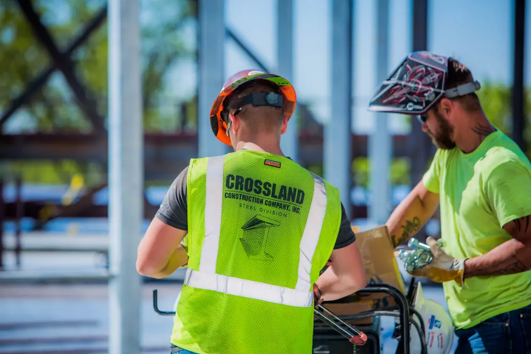 Two construction workers in safety gear work at a job site. One, wearing a vest labeled "Crossland Construction Company, Inc.," faces away, while the other sorts materials. Steel beams are visible in the background.