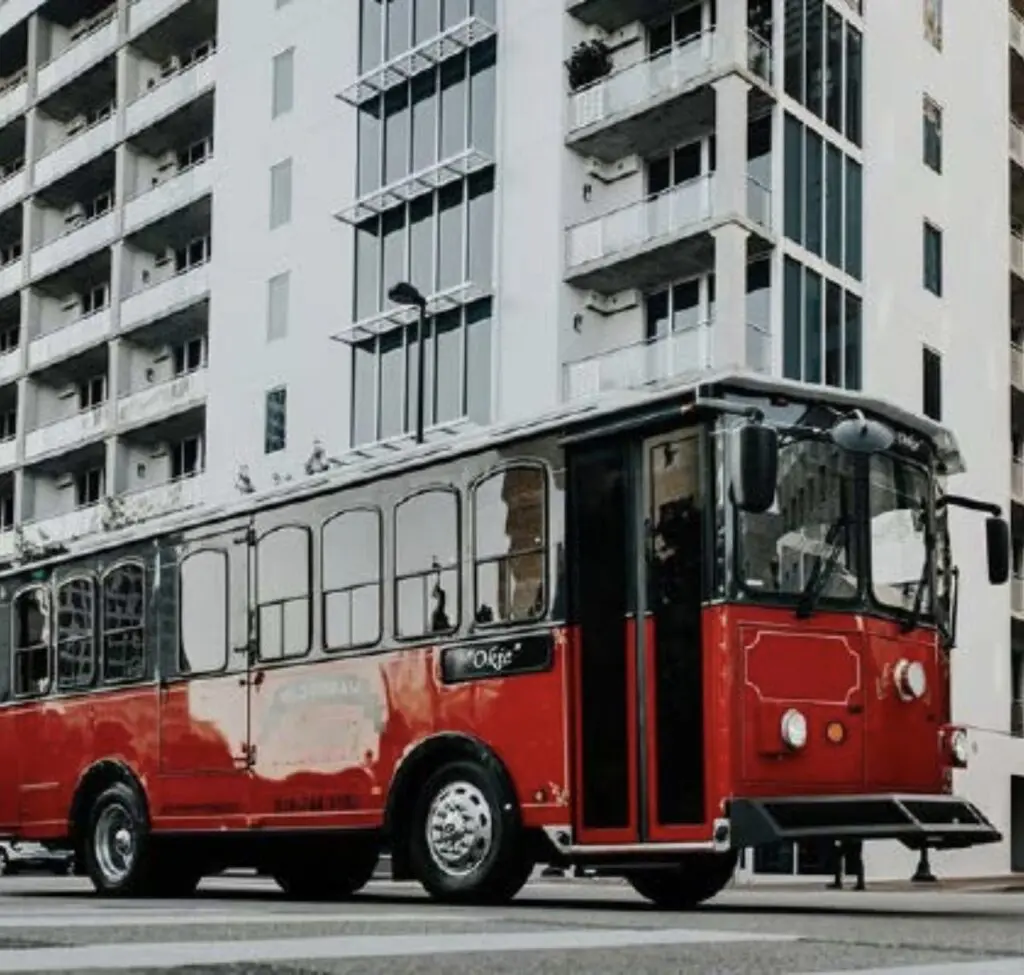 A red vintage-style trolley bus is parked on a city street in front of a modern multi-story apartment building.