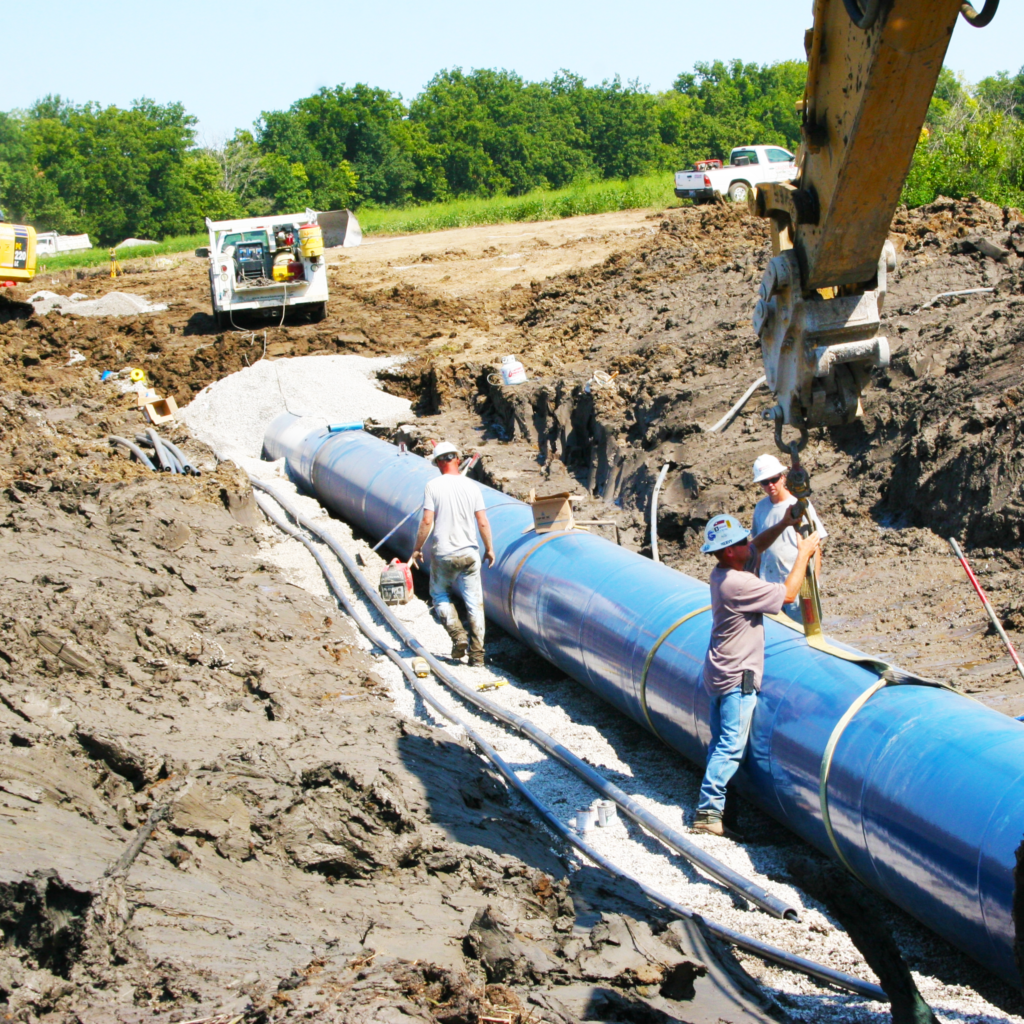 Construction workers install a large blue pipeline in a muddy trench, using heavy machinery and surrounded by construction vehicles and trees in the background.