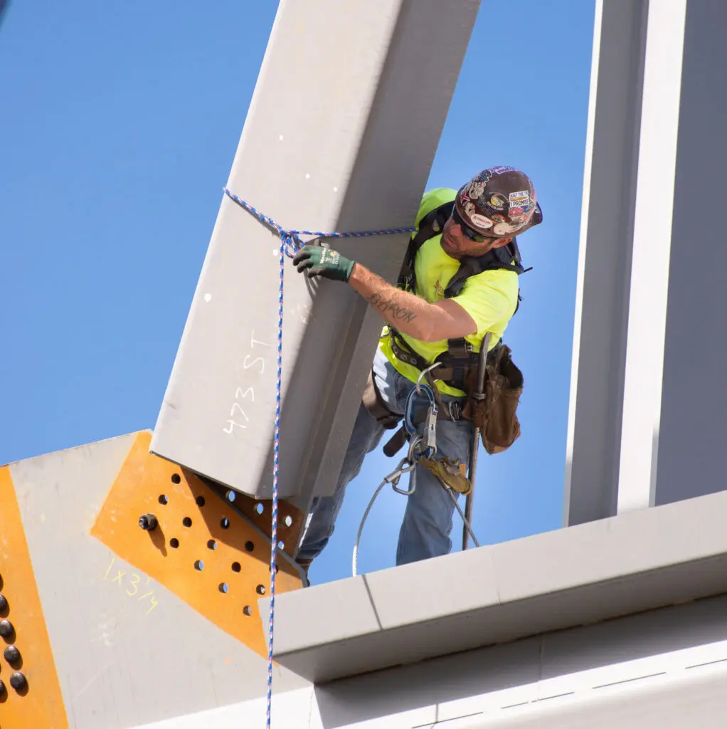 A construction worker in safety gear secures a large steel beam with a rope while standing on a structure, against a clear blue sky.