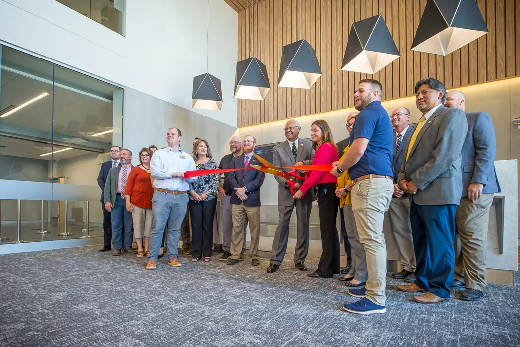A group of people in business attire stand together indoors, with two individuals holding oversized scissors to cut a red ribbon, suggesting a ribbon-cutting ceremony in a modern office setting.