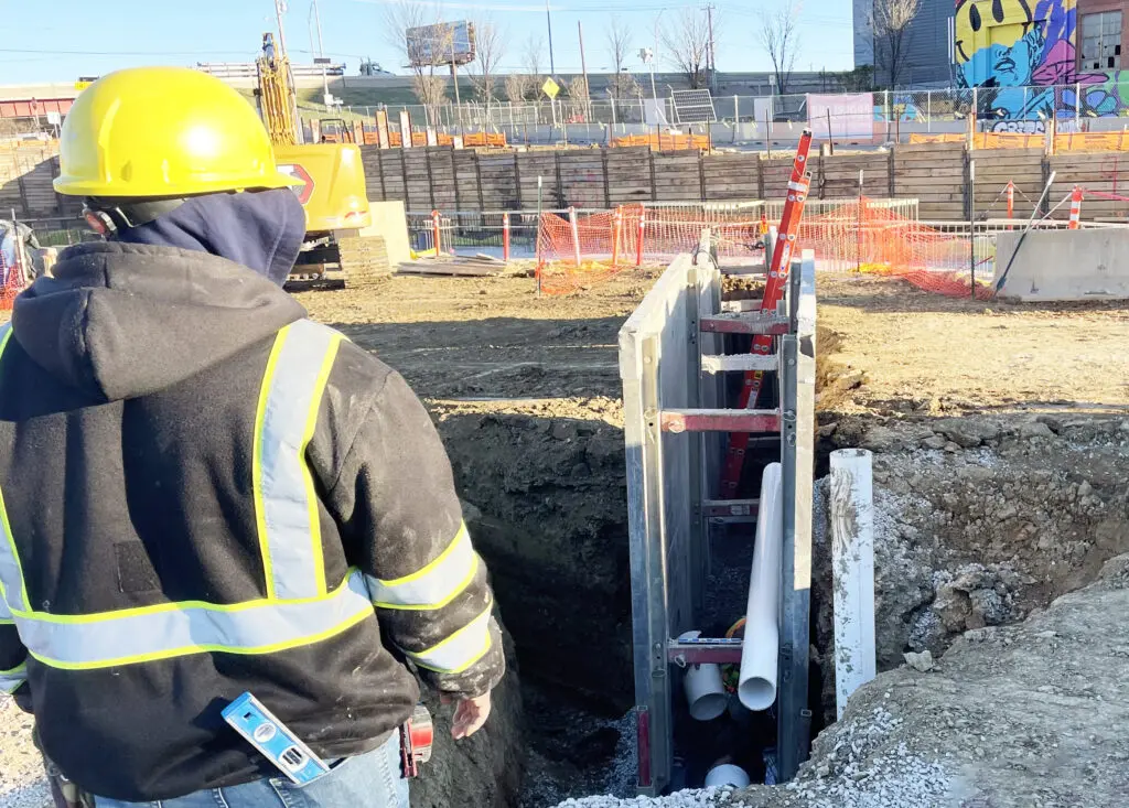 A construction worker in a yellow hard hat and safety jacket stands beside a trench with pipes and a trench box at a construction site, with equipment and safety barriers in the background.