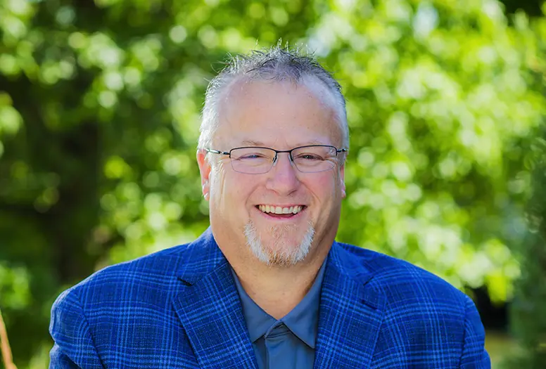 A middle-aged man with short gray hair, glasses, and a goatee smiles outdoors. He is wearing a blue plaid blazer over a gray shirt, with a blurred background of green foliage.