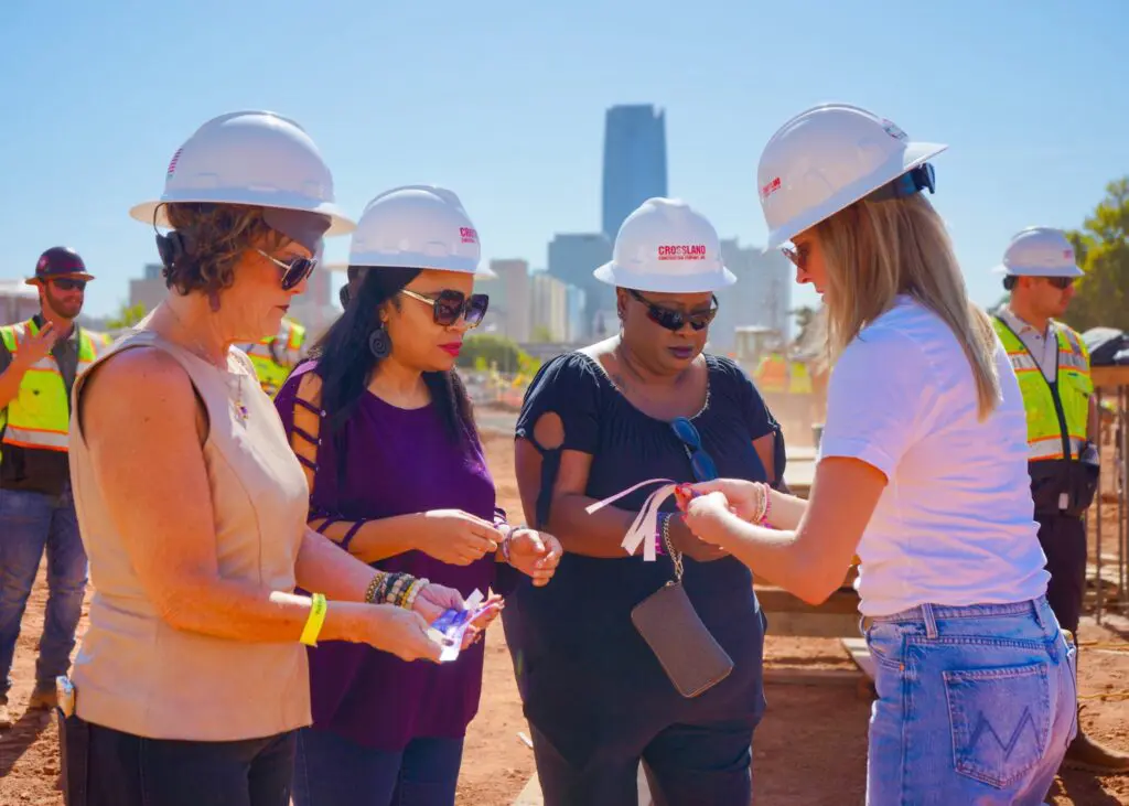 Four women wearing white construction helmets stand together on a construction site, holding ribbons. Two construction workers and a city skyline are visible in the background under a clear blue sky.