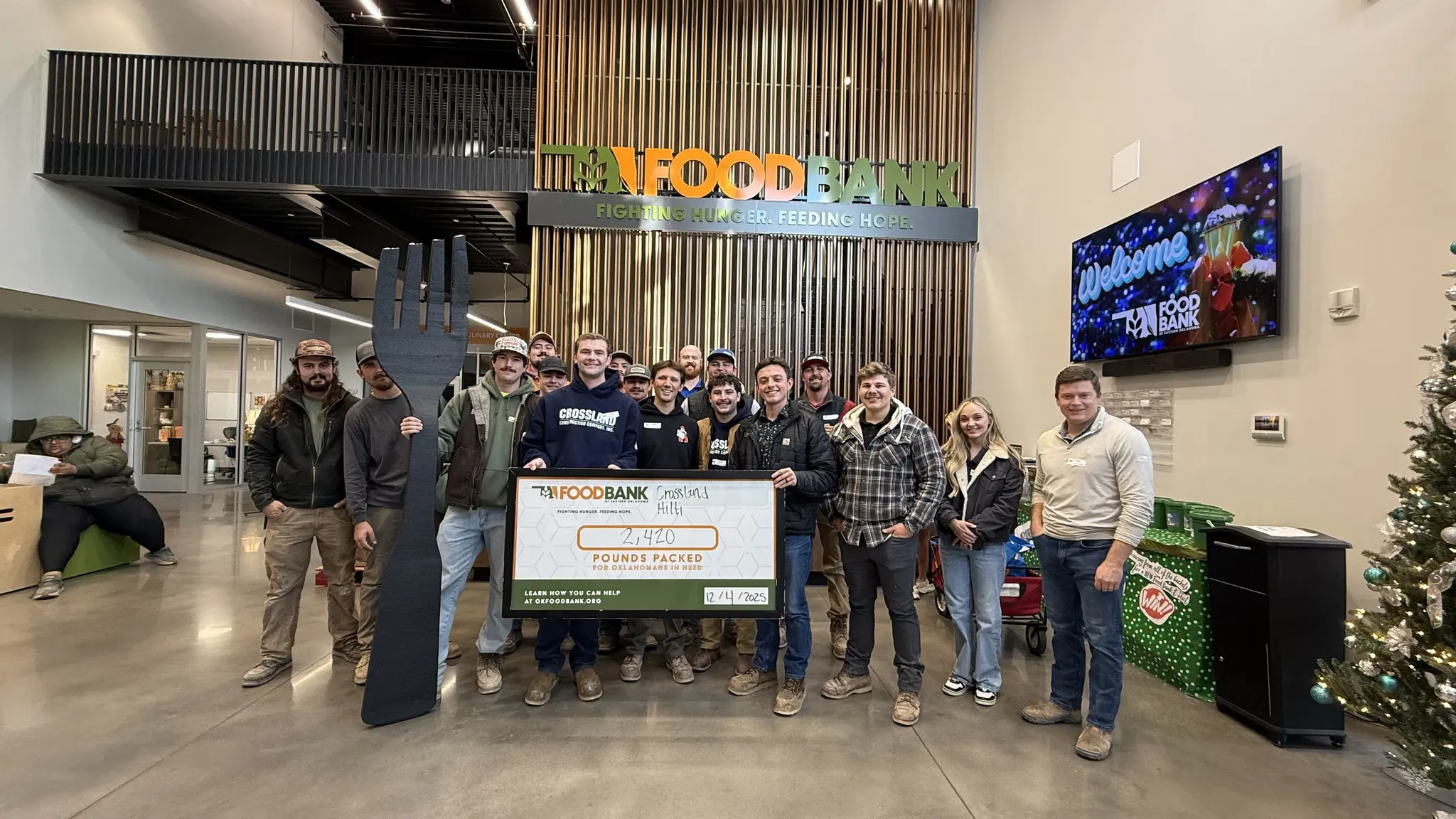 A group of people pose inside a food bank, holding a large check displaying "4,440 Pounds Packed" and a giant black fork. The food bank sign and a holiday tree are visible in the background.