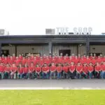 A large group of people wearing matching red shirts and jeans pose for a group photo in front of a building labeled “THE SHOP.” The group is arranged in rows on a paved area with green grass in the foreground.