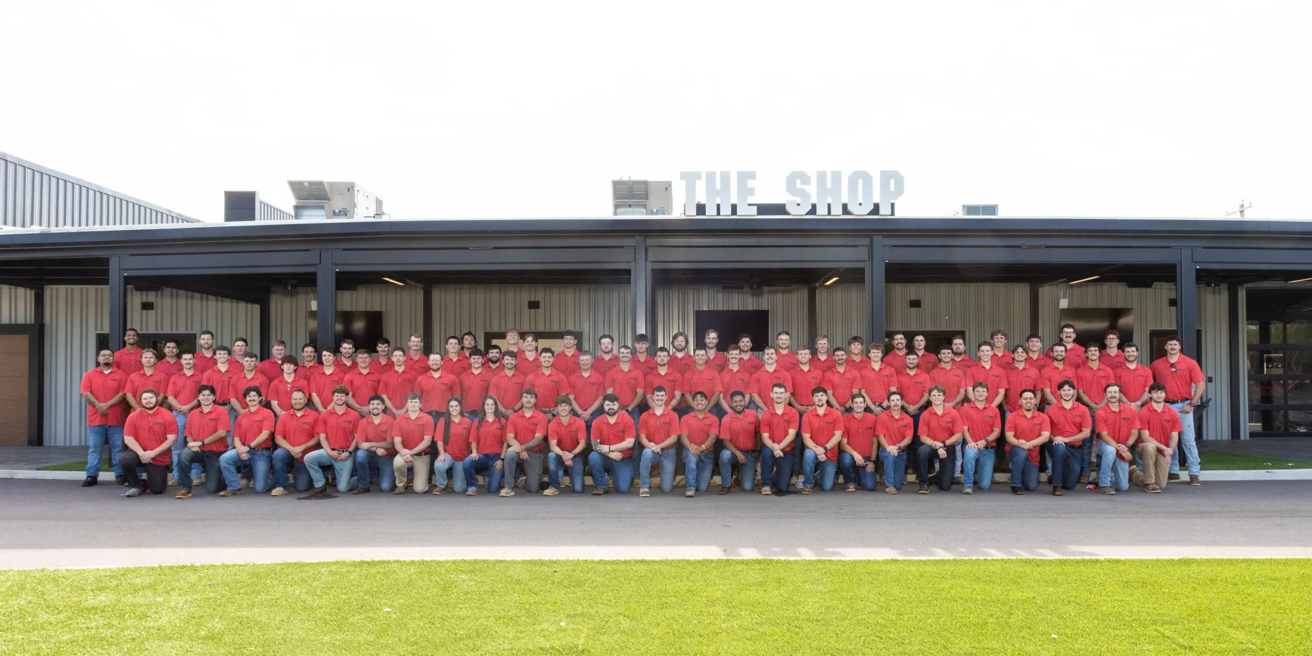 A large group of people wearing matching red shirts and jeans pose for a group photo in front of a building labeled “THE SHOP.” The group is arranged in rows on a paved area with green grass in the foreground.