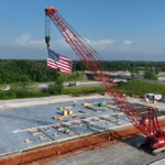 A large red construction crane with an American flag hanging from its hook sits on a building site with concrete foundation, surrounded by greenery and a highway under a clear blue sky.