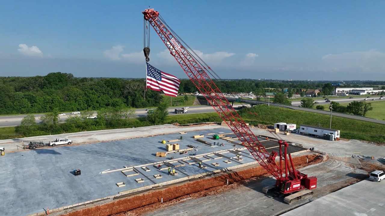 A large red crane with an American flag hanging from its hook is positioned on a construction site with a concrete foundation, surrounded by greenery and a highway in the background.