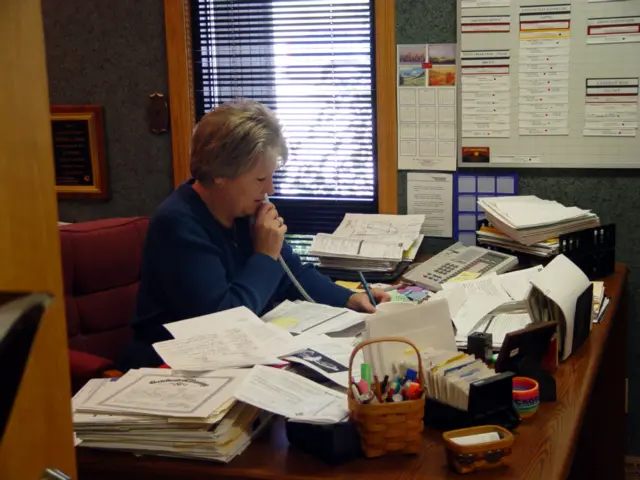 A woman sits at a cluttered office desk, surrounded by papers, folders, and office supplies. She appears to be thinking or reading, holding a pen to her mouth, with a window and a wall calendar in the background.