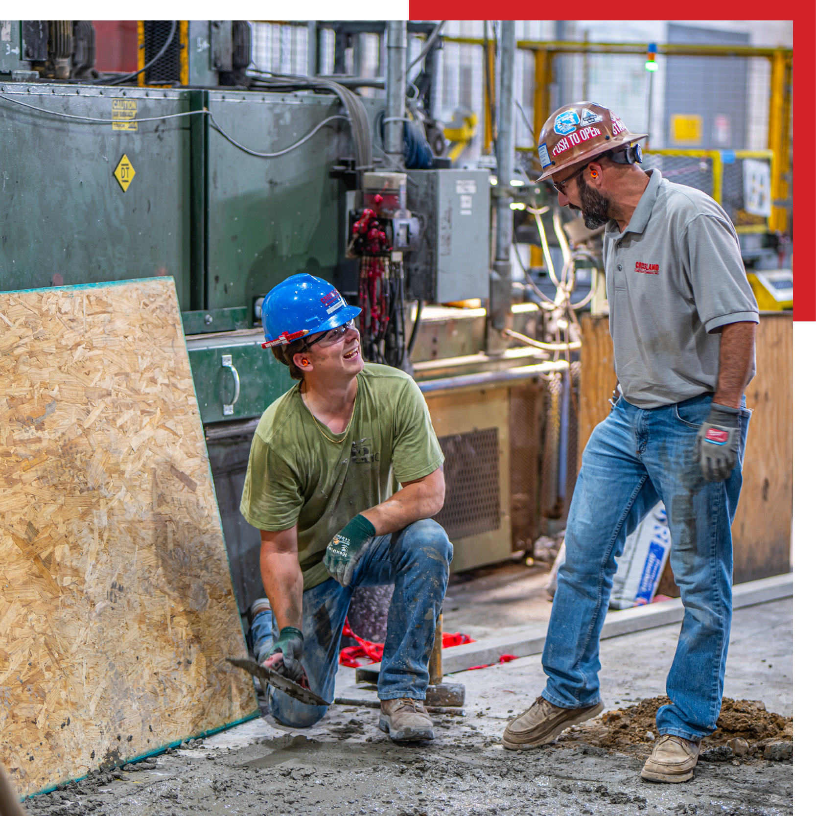 Two construction workers wearing hard hats and gloves are working with wet concrete indoors, near industrial equipment. One is kneeling and using a trowel, while the other stands talking to him.