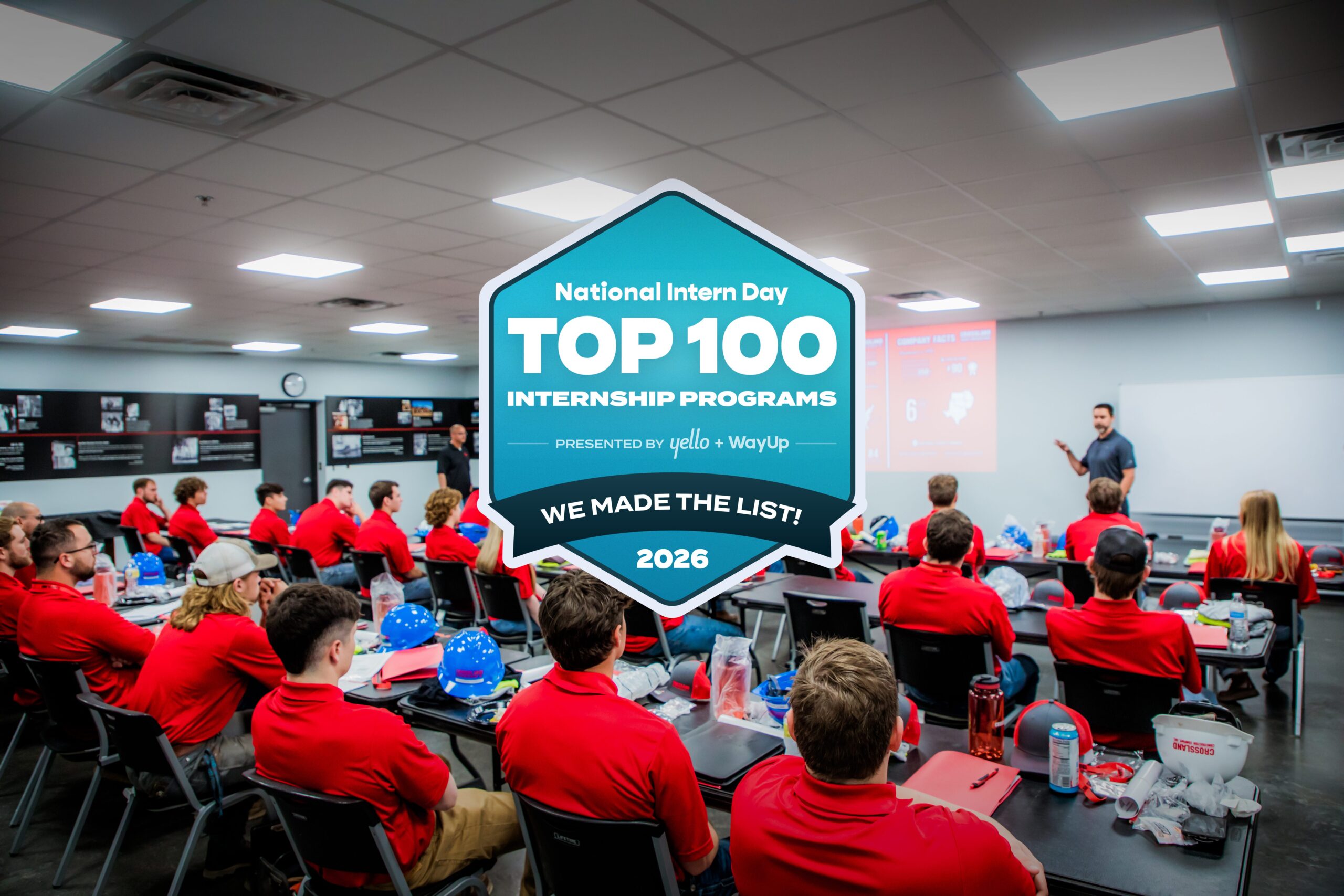 A group of interns in red shirts listen to a presenter in a classroom. A large badge in the center reads “Top 100 Internship Programs, 2026, National Intern Day.”.