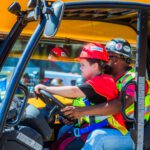 A child wearing a red hard hat and safety vest sits at the wheel of a construction vehicle, guided by an adult in similar safety gear, with a yellow machine in the background.