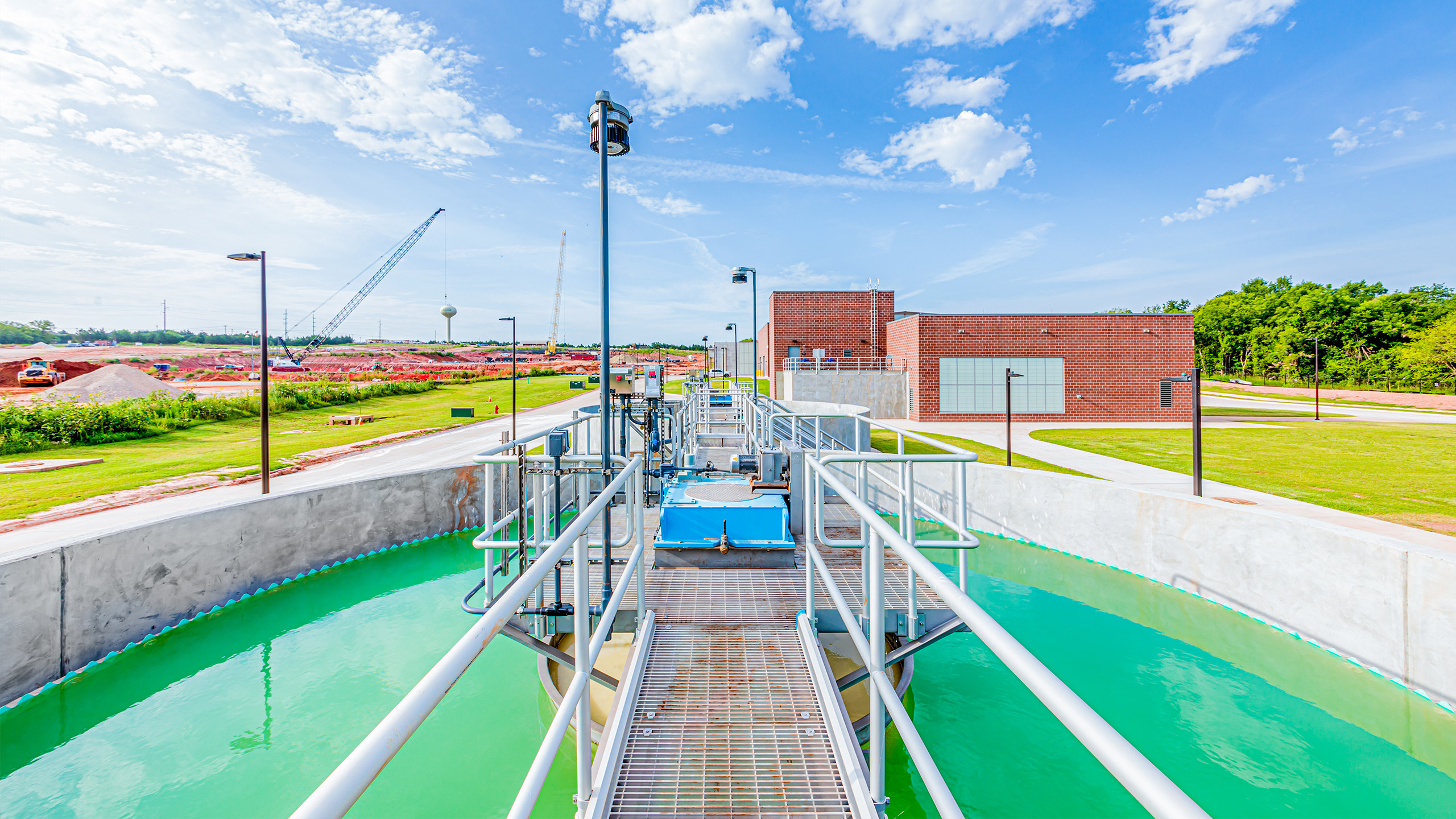 A water treatment plant with metal walkways over a circular tank of greenish water, surrounded by grass, red soil, and a brick building under a blue sky with scattered clouds.