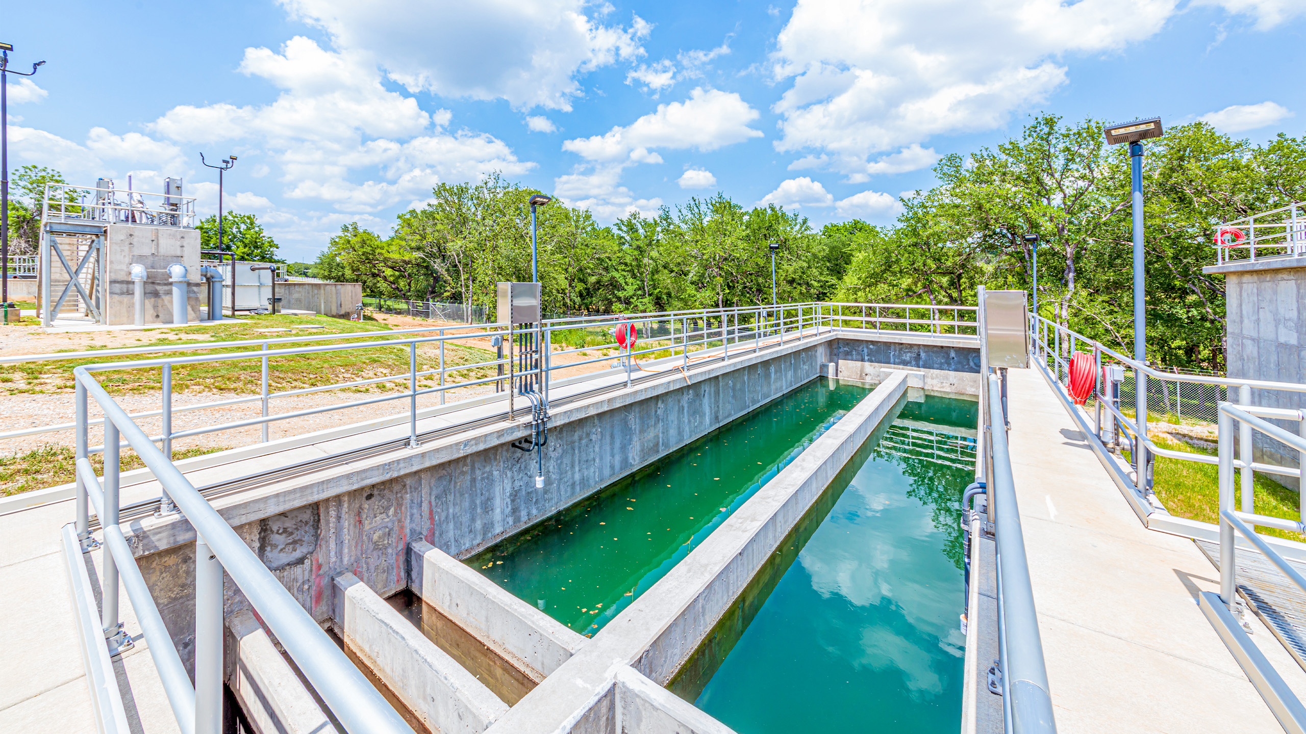 A water treatment facility with clear water flowing through open concrete channels, surrounded by metal railings and trees under a bright, partly cloudy sky.
