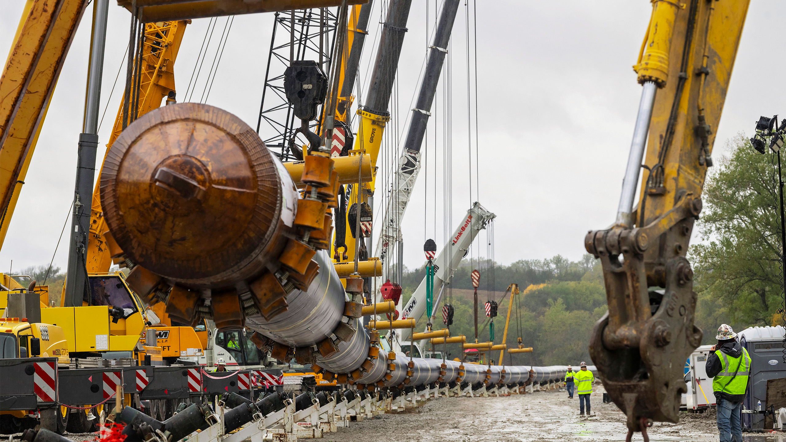 Large construction machinery lifts and aligns a long metal pipeline outdoors. Workers in safety gear stand nearby. The scene is muddy and overcast, with trees in the background and several cranes and equipment in use.