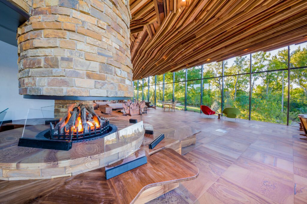 A modern lounge with large windows showing trees outside, a central stone fireplace with a burning fire, wooden floors, and uniquely designed, sloped wooden ceilings. Contemporary chairs are set near the windows.