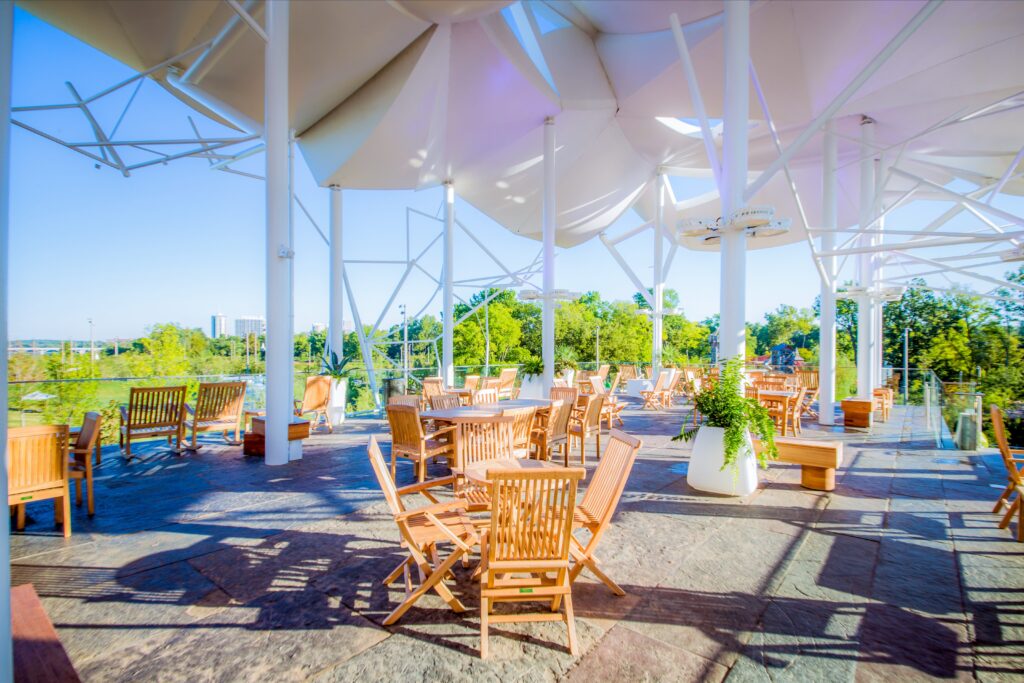A spacious outdoor patio with wooden tables and chairs sits under a modern white canopy structure. The area is surrounded by trees and greenery, with sunlight casting shadows on the stone floor.