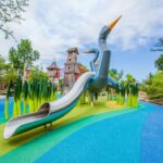A colorful playground features a large blue heron-themed slide surrounded by green cattail sculptures on a vibrant rubber surface, with whimsical wooden towers and trees in the background.