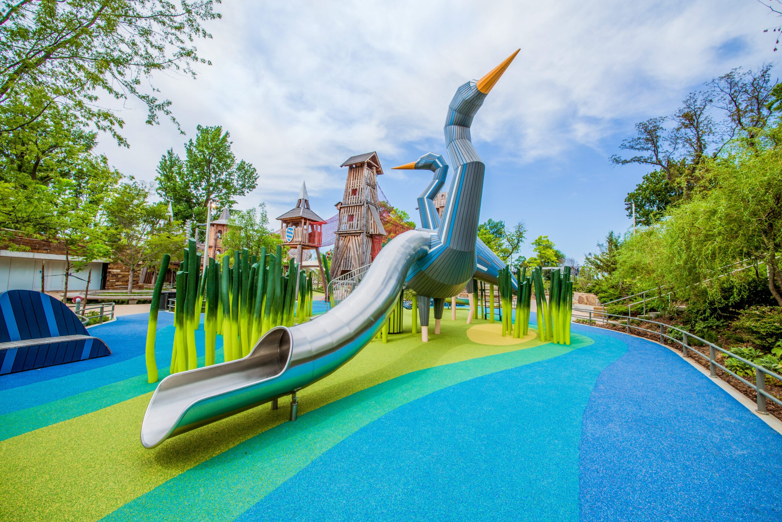 A colorful playground features a large blue heron-themed slide surrounded by green cattail sculptures on a vibrant rubber surface, with whimsical wooden towers and trees in the background.