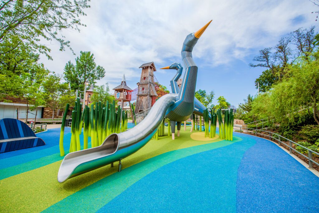A large playground slide shaped like two blue herons stands among tall green structures, on a colorful blue and green rubber surface, with trees and wooden towers in the background.