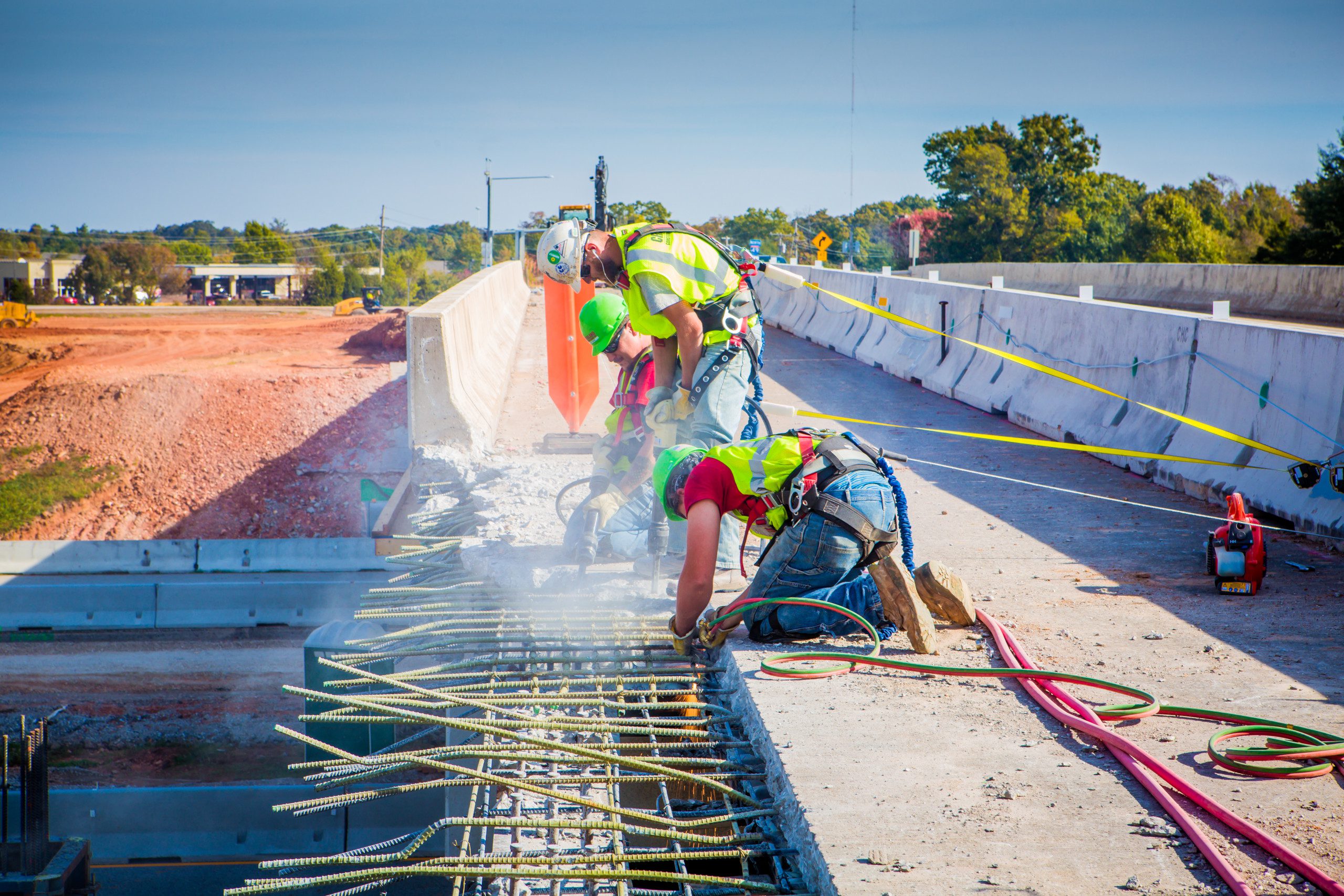 Construction workers in safety gear bend over rebar and concrete on a bridge under construction, using power tools with dust and debris visible; bright safety vests and helmets are worn.