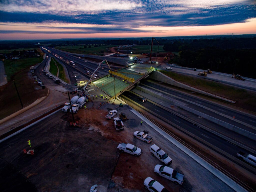 Aerial view of a highway bridge under construction at dusk, with workers, vehicles, and construction equipment on site. Concrete is being poured, and traffic flows on adjacent lanes under a dramatic, cloud-filled sky.