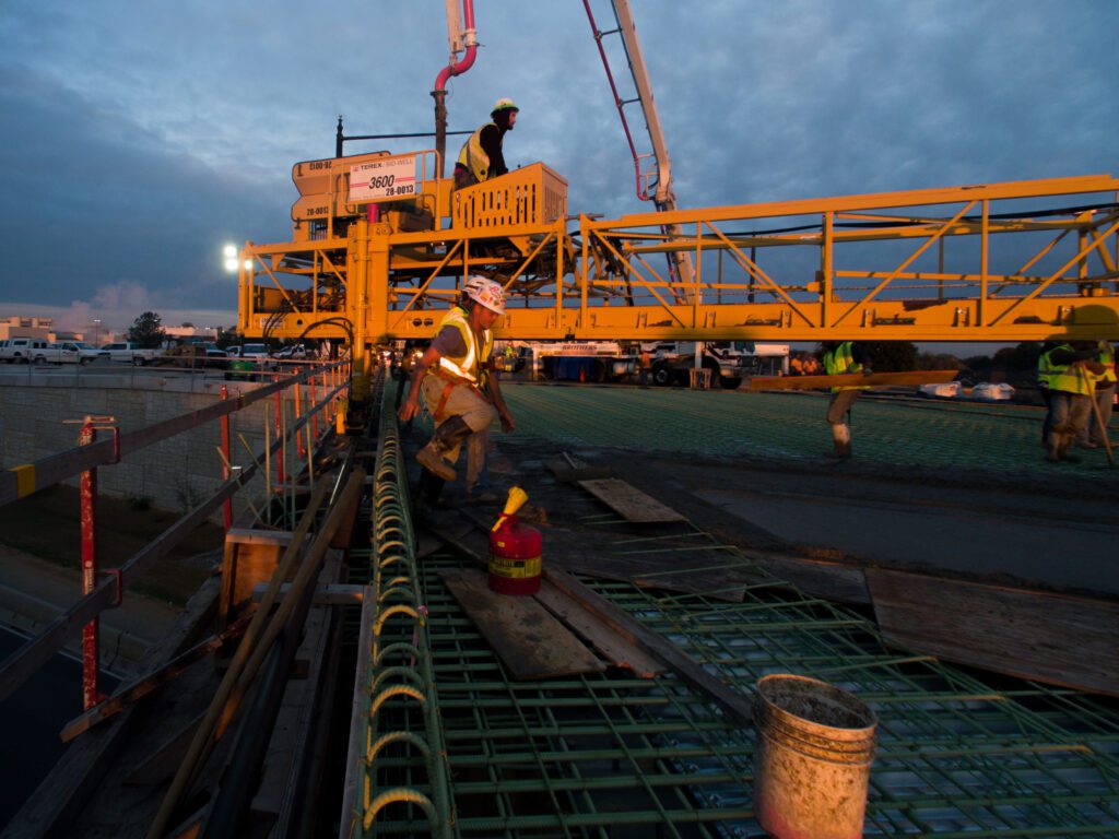 Construction workers in safety gear work on a bridge at dusk, with a large yellow cement pump machine overhead and green rebar laid out on the structure. The sky is cloudy and illuminated by construction lights.