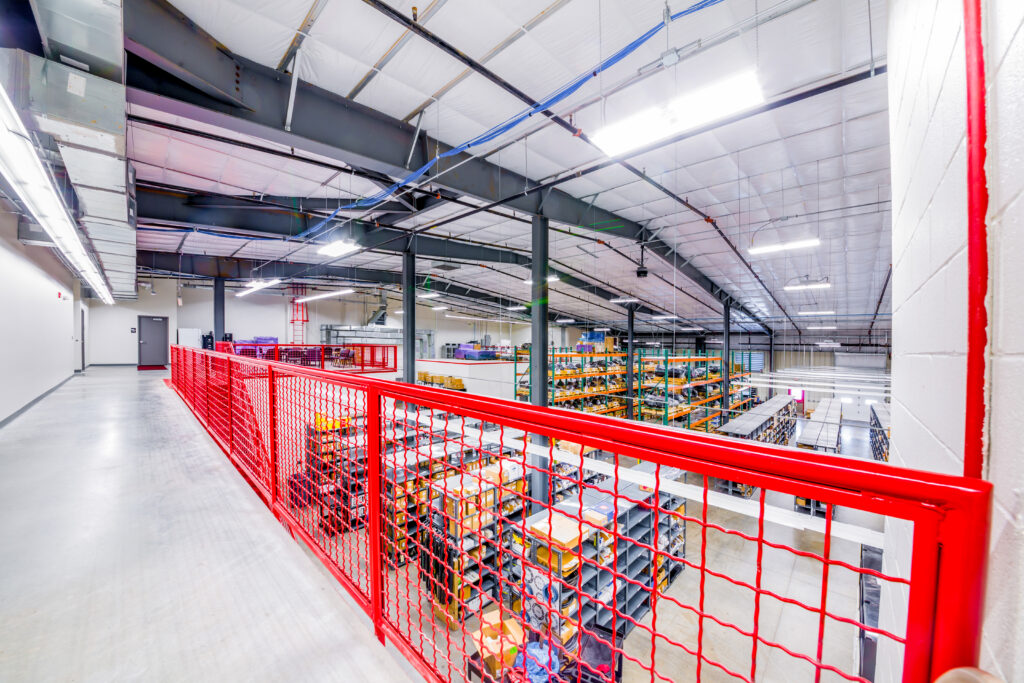 View from an upper walkway with red railing overlooking a large warehouse filled with organized shelves stocked with boxes and goods, under bright lights and a high, industrial ceiling.