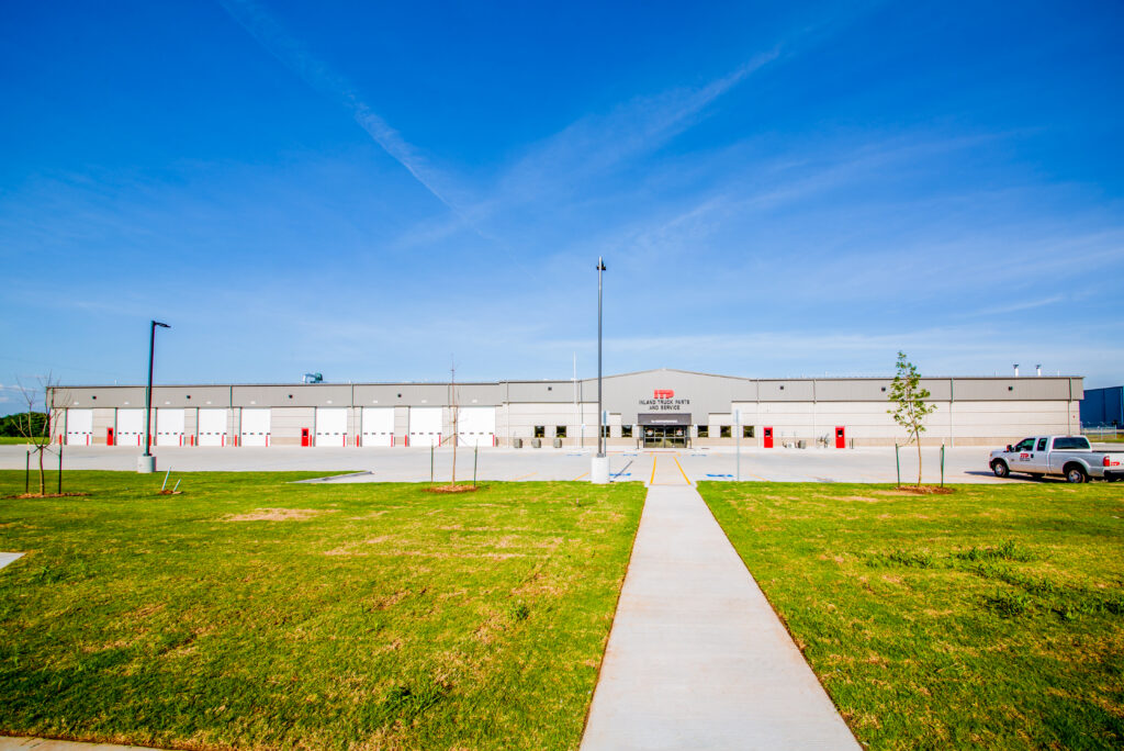 A wide, single-story industrial warehouse with many white bay doors, a few red doors, a clear blue sky, and a large, empty grassy area in front with a concrete walkway leading to the entrance.