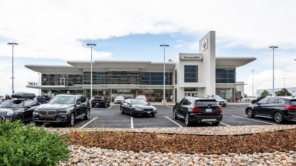 A modern BMW dealership with large glass windows, a "Grand Reopening" sign, and several parked cars in front. The sky is partly cloudy and landscaping with rocks and mulch is visible in the foreground.