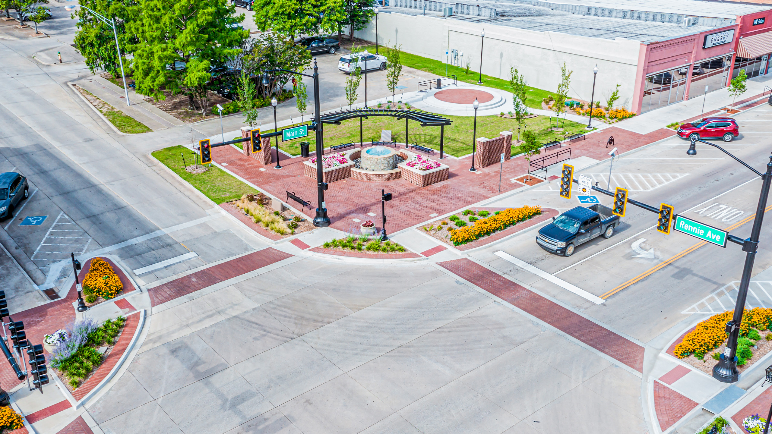Aerial view of an intersection with cars, traffic lights, crosswalks, landscaped flower beds, and a small park area with a circular brick feature and benches. Nearby buildings and parked cars are visible.