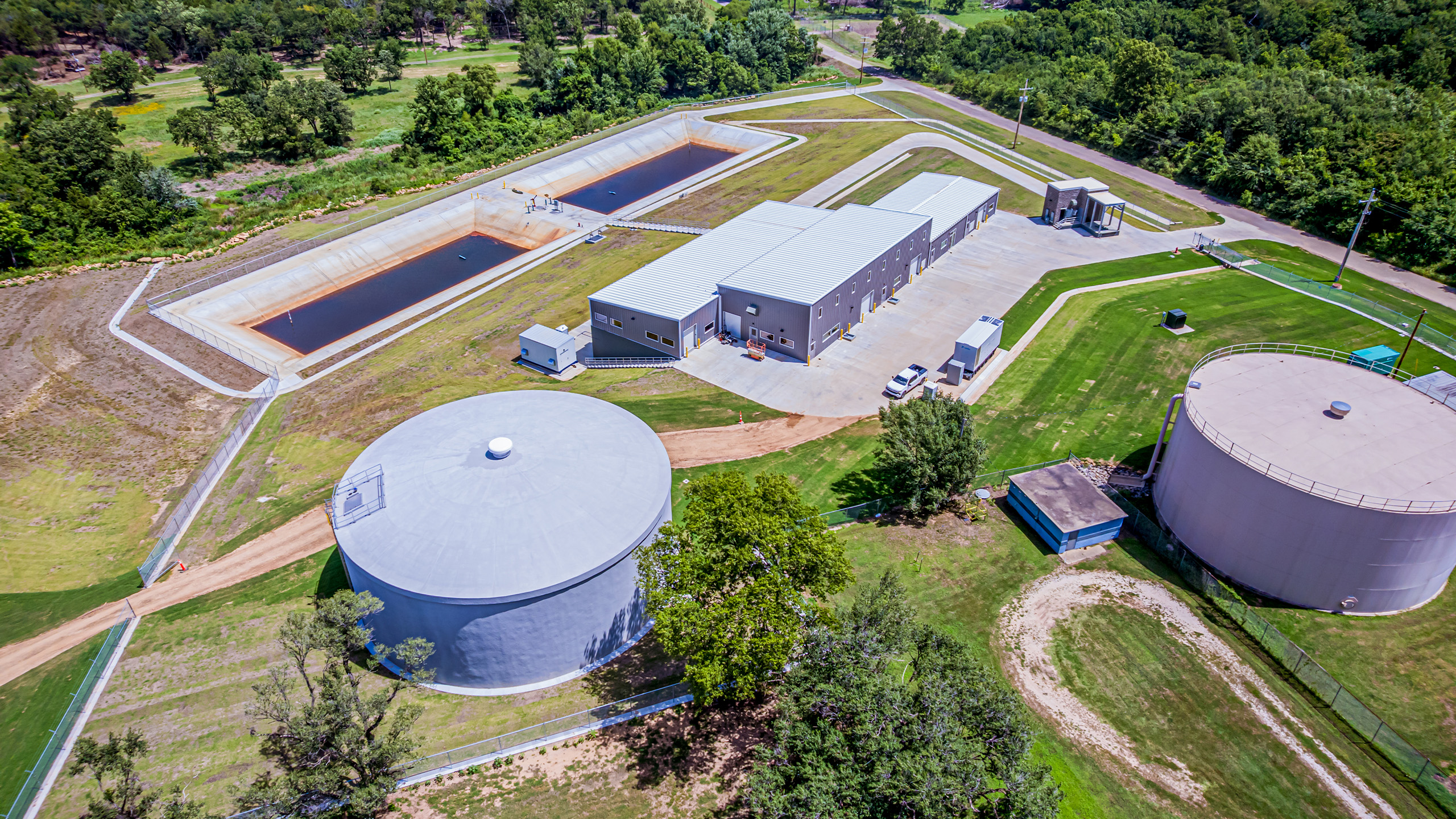 Aerial view of a water treatment plant with large storage tanks, rectangular water basins, and industrial buildings surrounded by green grass and trees.