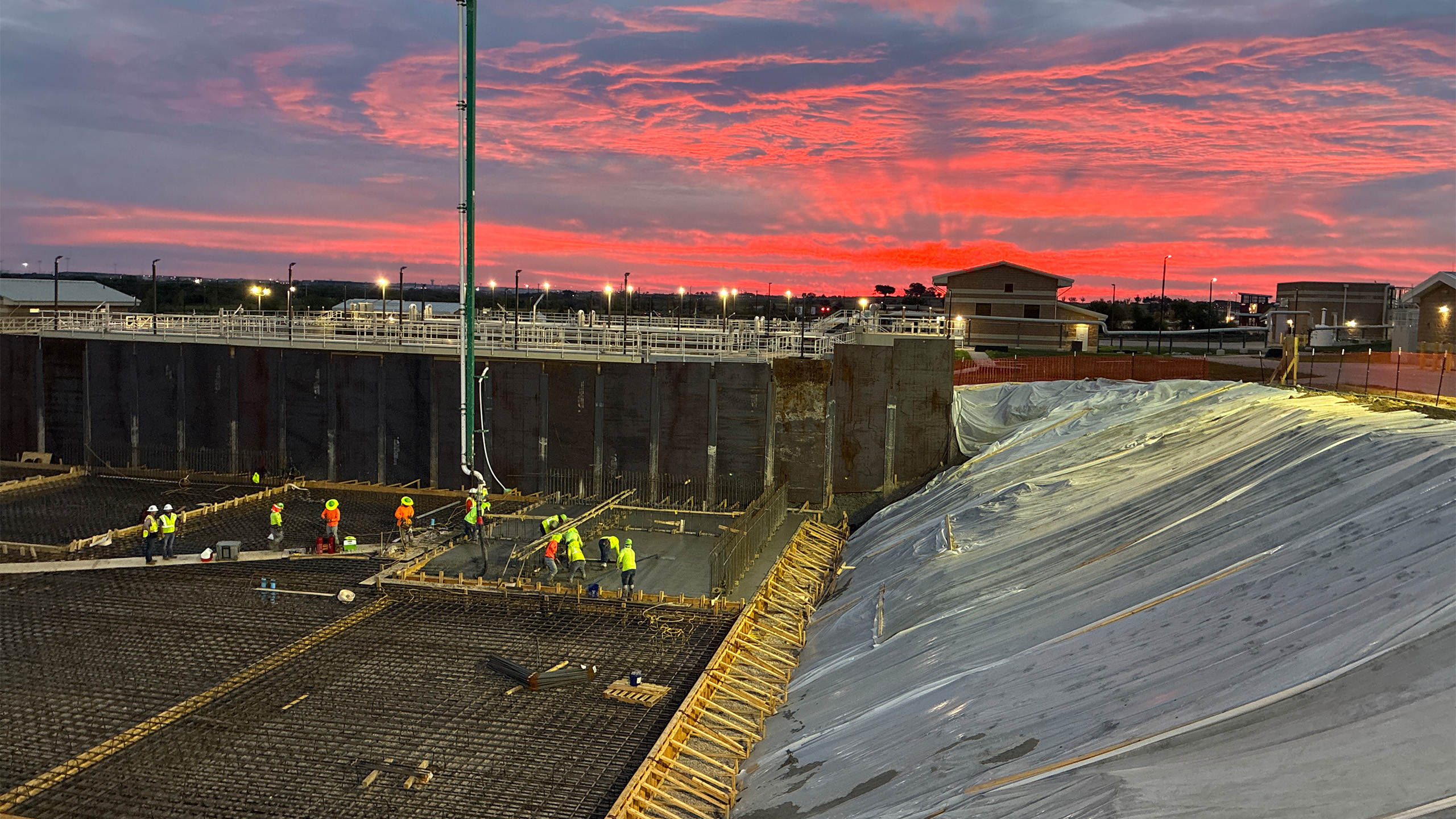 Construction workers in safety gear work on a large foundation site at sunset, with steel rebar on the ground and a vivid red sky in the background. Plastic sheets cover a sloped section on the right side of the image.