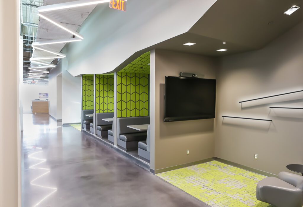 Modern office space with gray and green hexagon-patterned booth seating, a large wall-mounted TV, geometric ceiling lights, and a polished concrete floor. The area has a contemporary, open design.