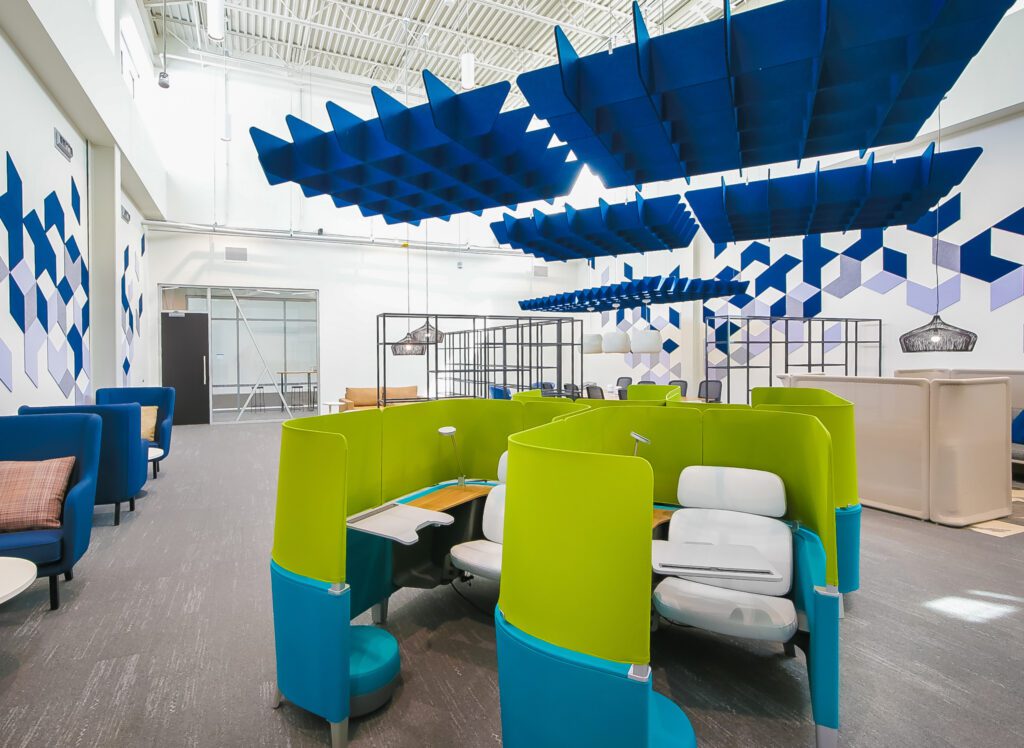 A modern office space with bright green and blue curved workstations, geometric wall art, blue acoustic ceiling panels, and a high ceiling with abundant natural light.
