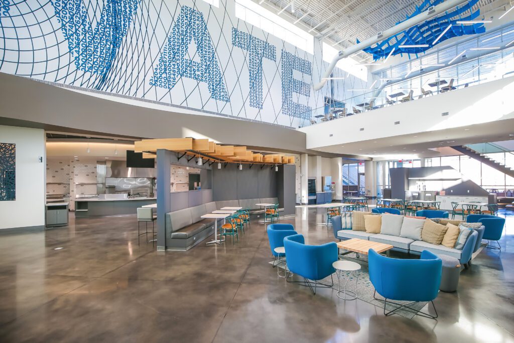 Modern, open office lounge with blue and tan seating, small tables, a kitchen area, and large windows. A mezzanine level and geometric wall art reading "ATE" are visible above.