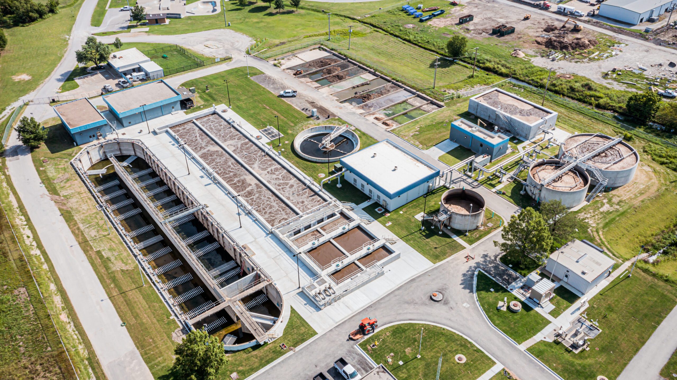 Aerial view of a large water treatment plant with multiple rectangular and circular tanks, blue-roofed buildings, surrounding roads, and green grassy areas.