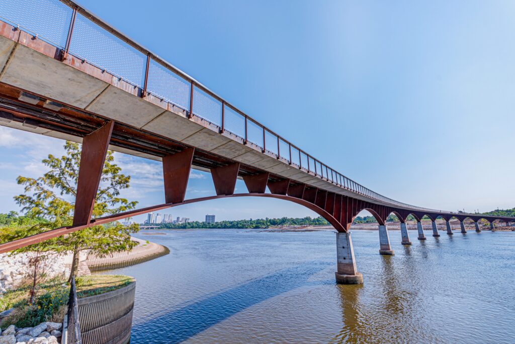 A long pedestrian bridge with rust-colored arches spans a wide river under a clear blue sky, with trees and city buildings visible in the background.