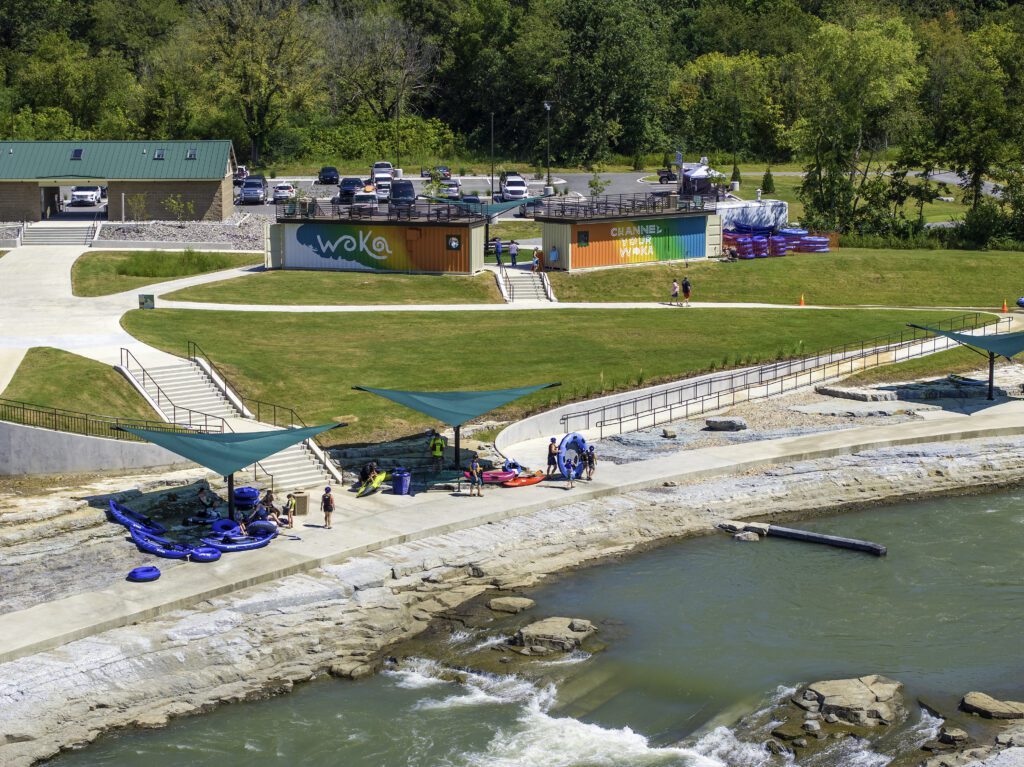 A riverside recreation area features grassy lawns, a modern building with "WOKA" signage, parked cars, and people preparing for water activities near blue rafts and sun shades by a flowing river.