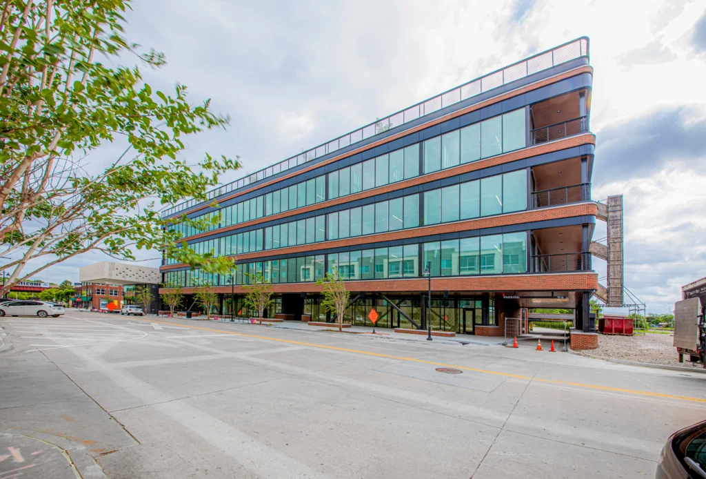 A modern, four-story building with large glass windows and brick accents stands on a quiet, empty street under a cloudy sky, with some trees and construction cones visible nearby.