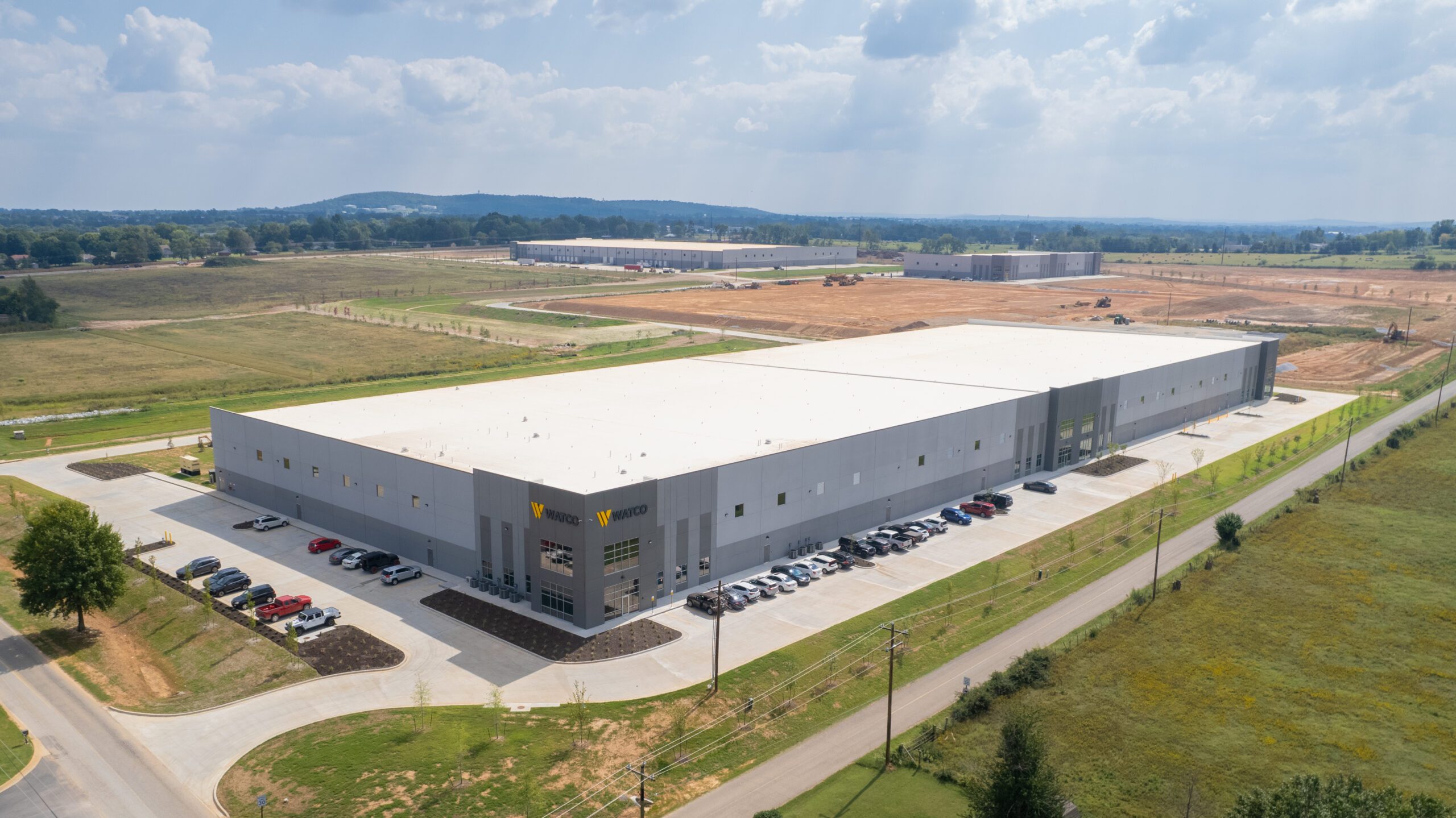 A large, rectangular warehouse with a white roof sits in a grassy, semi-rural area. Several cars are parked along the side, and more industrial buildings are visible in the distance under a partly cloudy sky.