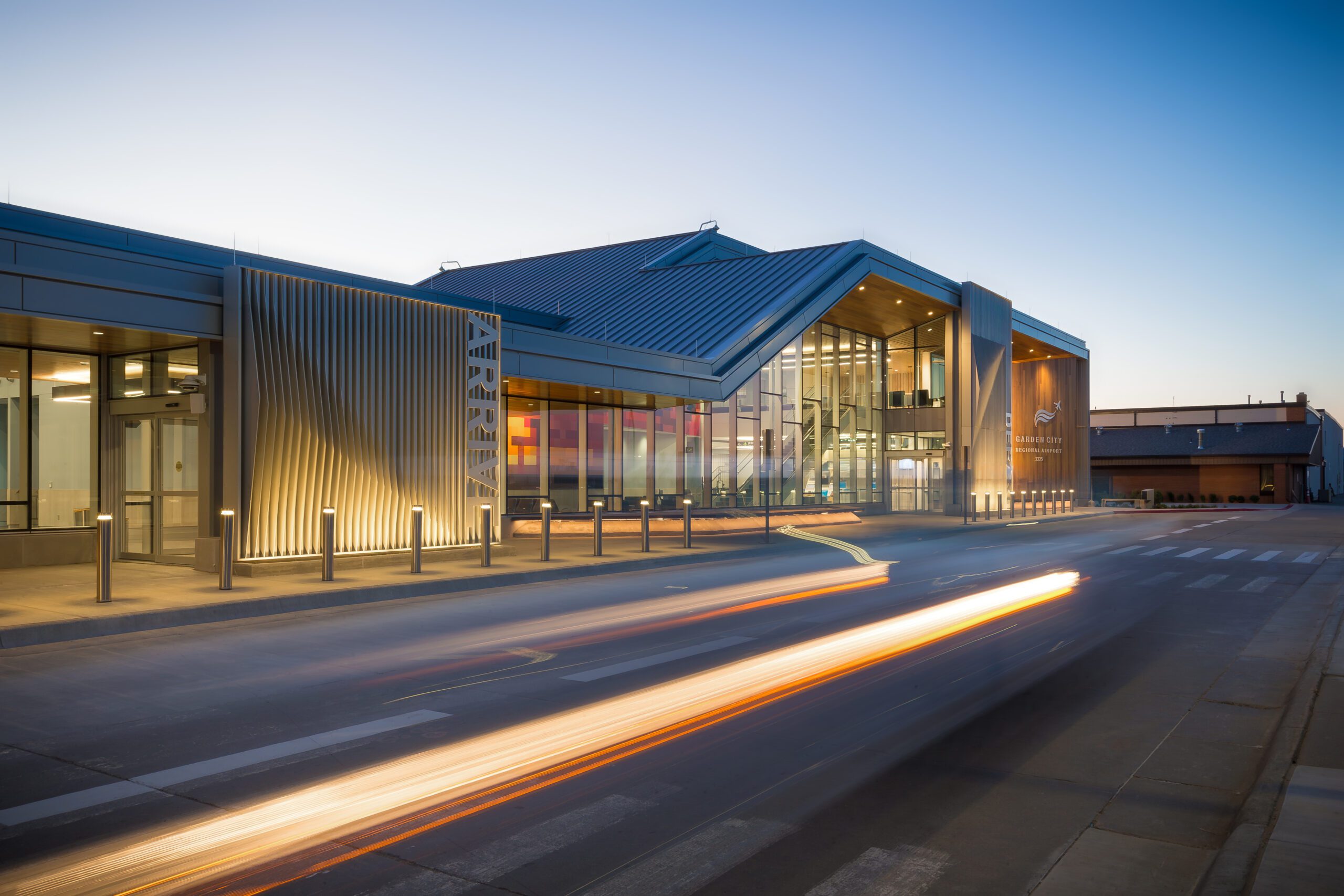 A modern airport terminal at dusk with glass walls and a zigzag roof, illuminated by warm lights. A car's headlights create streaks of light on the road in front.