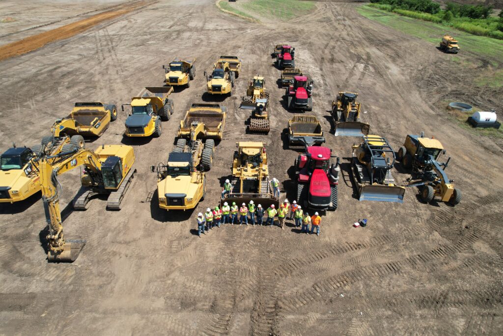 A group of construction workers in safety gear stand in front of an array of heavy machinery, including excavators, bulldozers, and trucks, on a large dirt lot under clear skies.