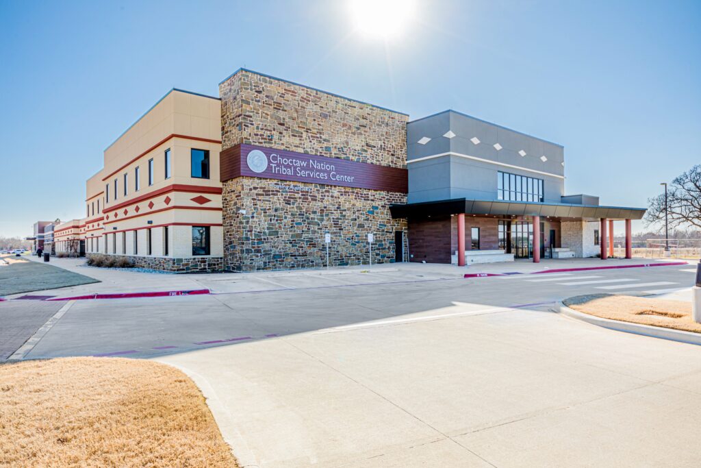 A modern building with stone and beige walls labeled "Choctaw Nation Tribal Services Center" sits under a clear blue sky, surrounded by a driveway and dry grass landscaping.