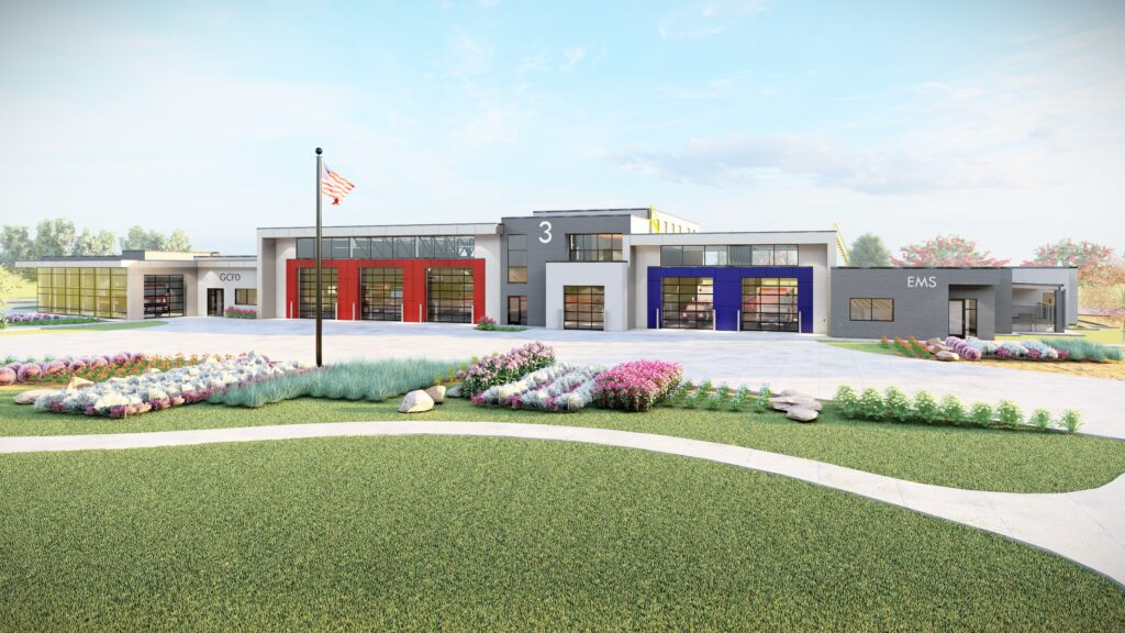 Modern fire and EMS station building with three colored garage doors (red, orange, blue), a flagpole, landscaped gardens, and a well-maintained lawn under a partly cloudy sky.