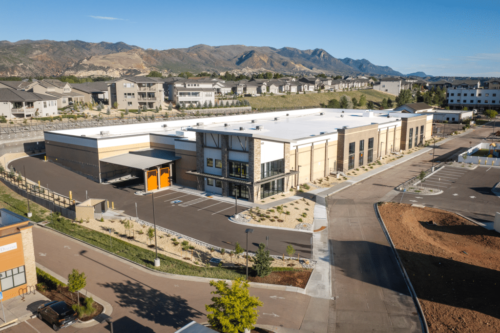 A large modern building with a white roof, surrounded by paved roads and landscaping, with neighborhoods and mountains in the background under a clear blue sky.
