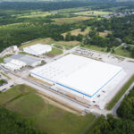 Aerial view of a large industrial facility with white rooftops, surrounded by green fields, trees, and nearby roads, with a small town visible in the background.