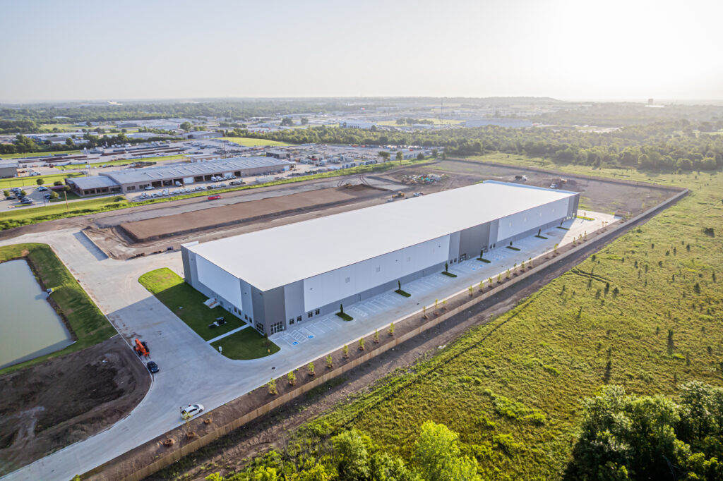Aerial view of a large, modern warehouse with a white roof, surrounded by parking lots, roads, green fields, and nearby industrial buildings under a clear sky.