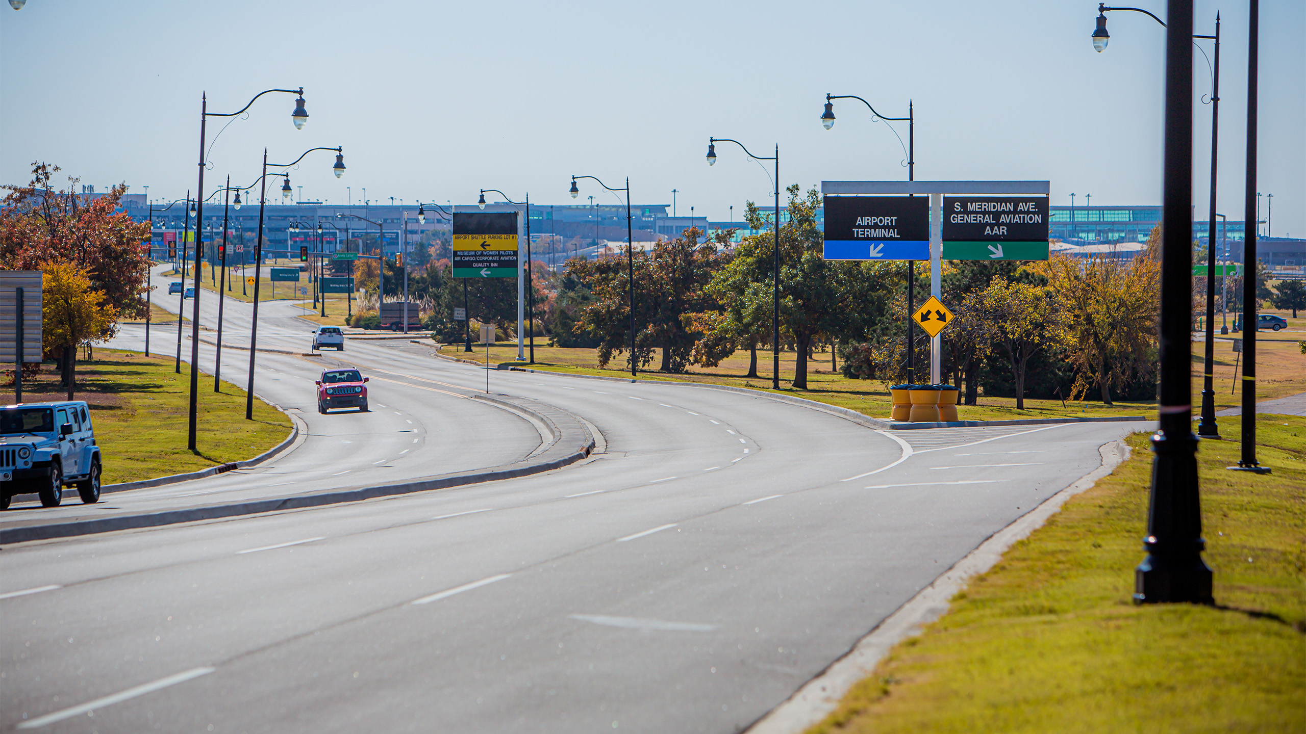 A wide road curves past streetlights and trees with cars driving. Overhead signs direct traffic toward the airport terminal and general aviation. The sky is clear and bright.